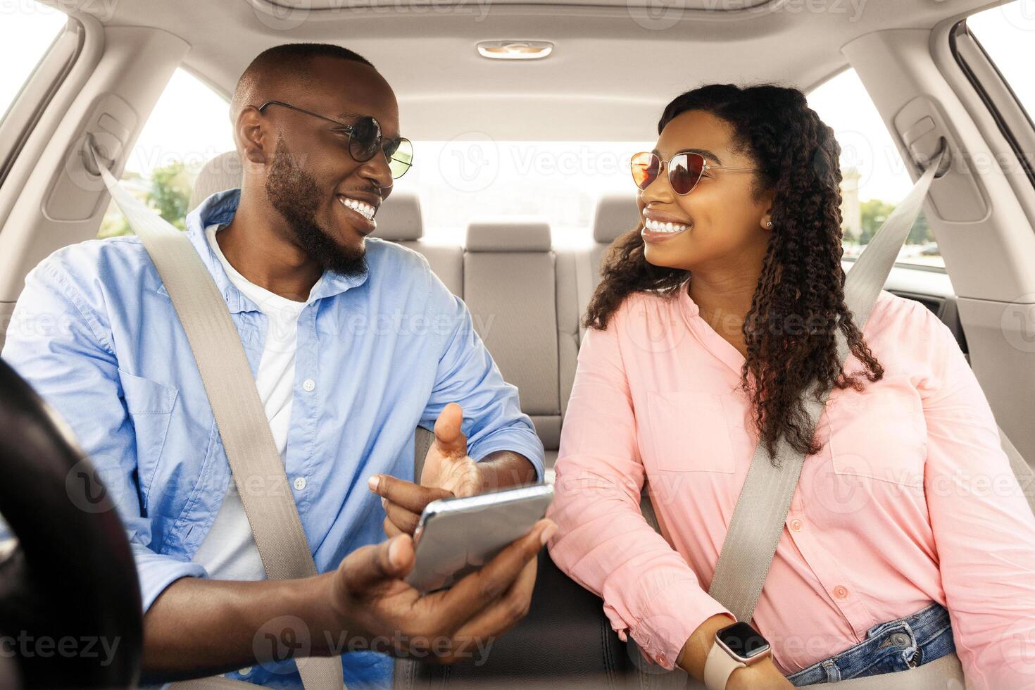 Vacation. Excited black male driver travelling with his beautiful girlfriend on new luxury automobile, showing shortcut way pointing at smartphone screen, couple in sunglasses looking at each other photo