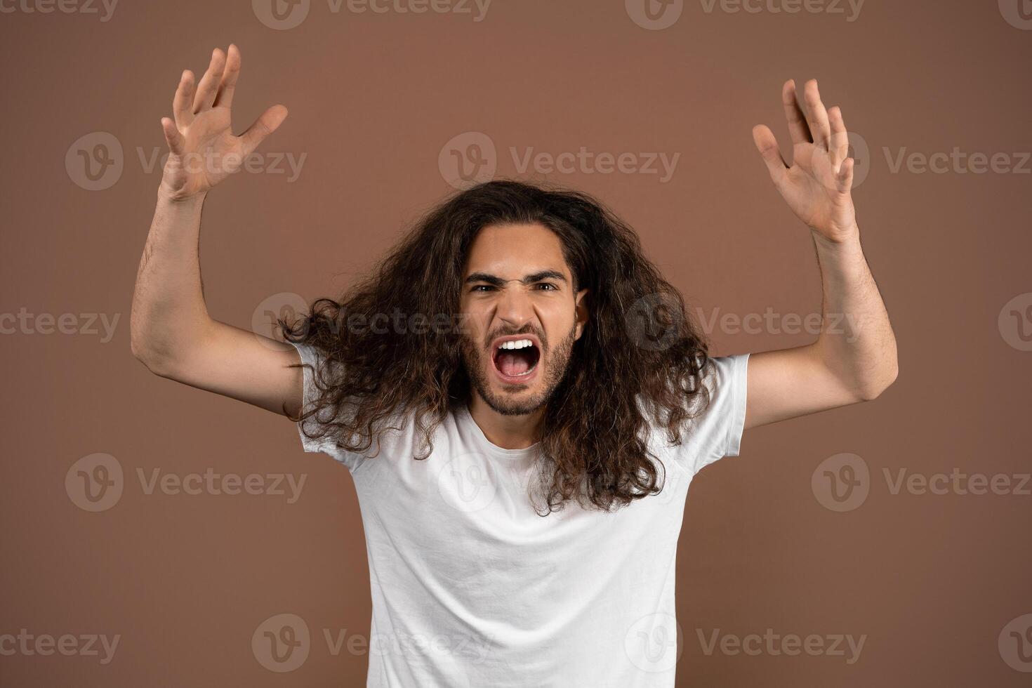 A young European man with long, curly hair displays strong emotions while standing against a plain brown background. His expressive pose and facial expression capture a moment of intensity. photo