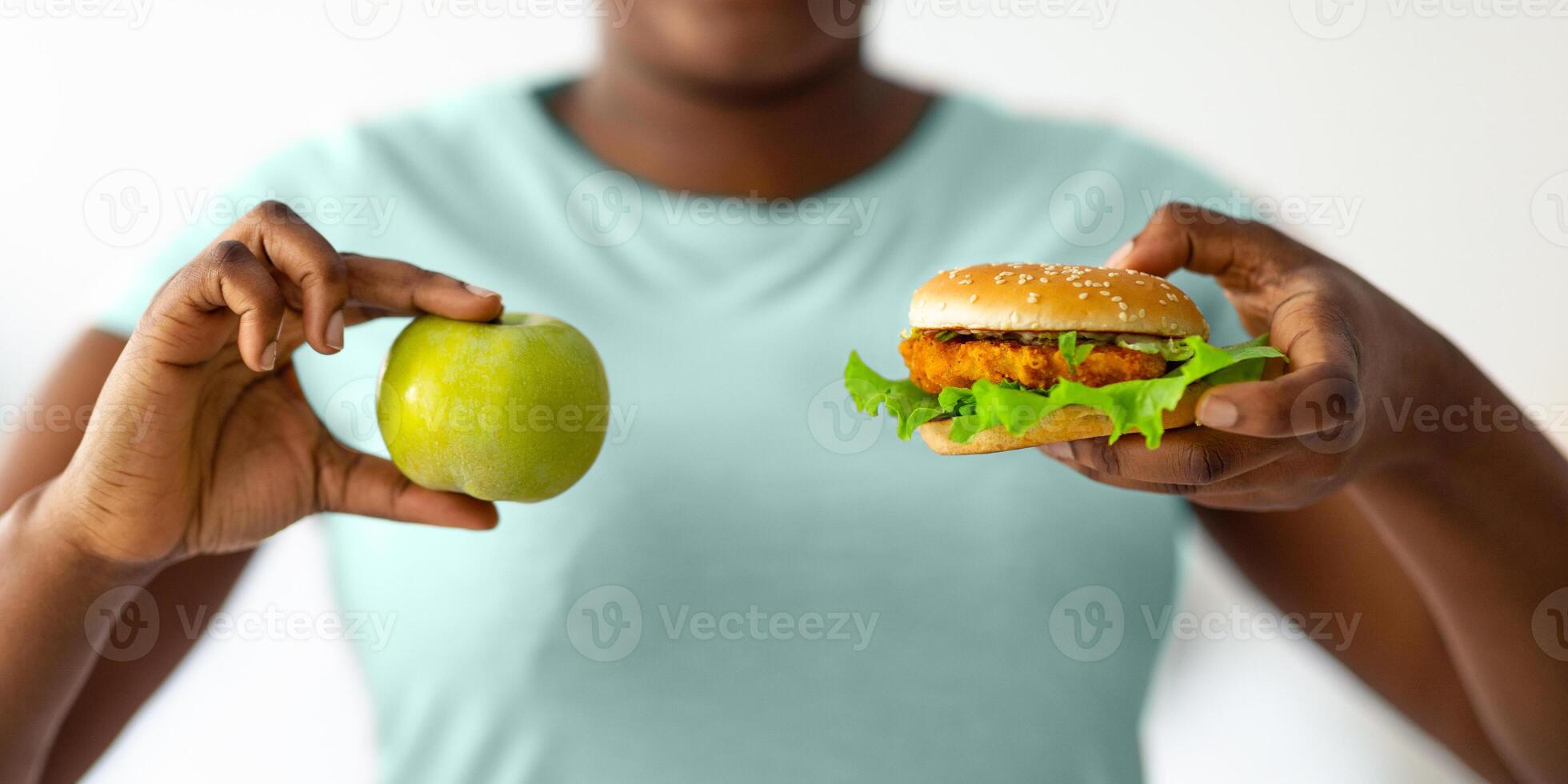 Unrecognizable overweight black woman choosing between burger and apple, closeup. Plus size young female on diet selecting between healthy food and junk meal. Nutrition for weight loss photo