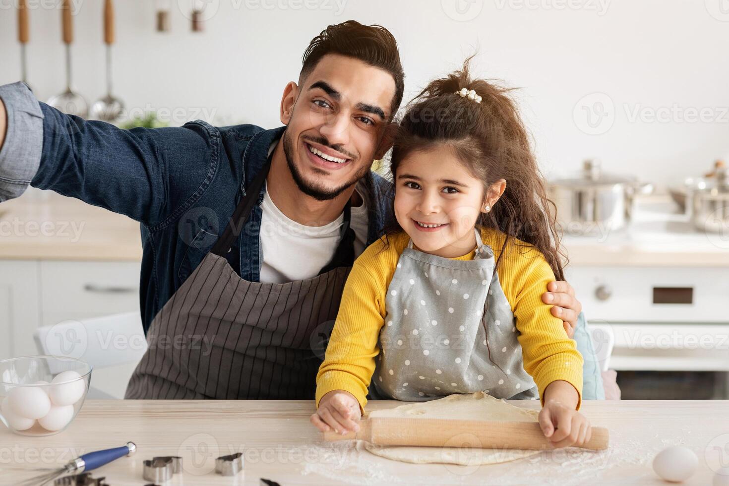 Happy young arab father and little daughter taking selfie in kitchen while baking, posing for camera while making homemade cookies together, cheerful middle eastern dad and child having fun at home photo