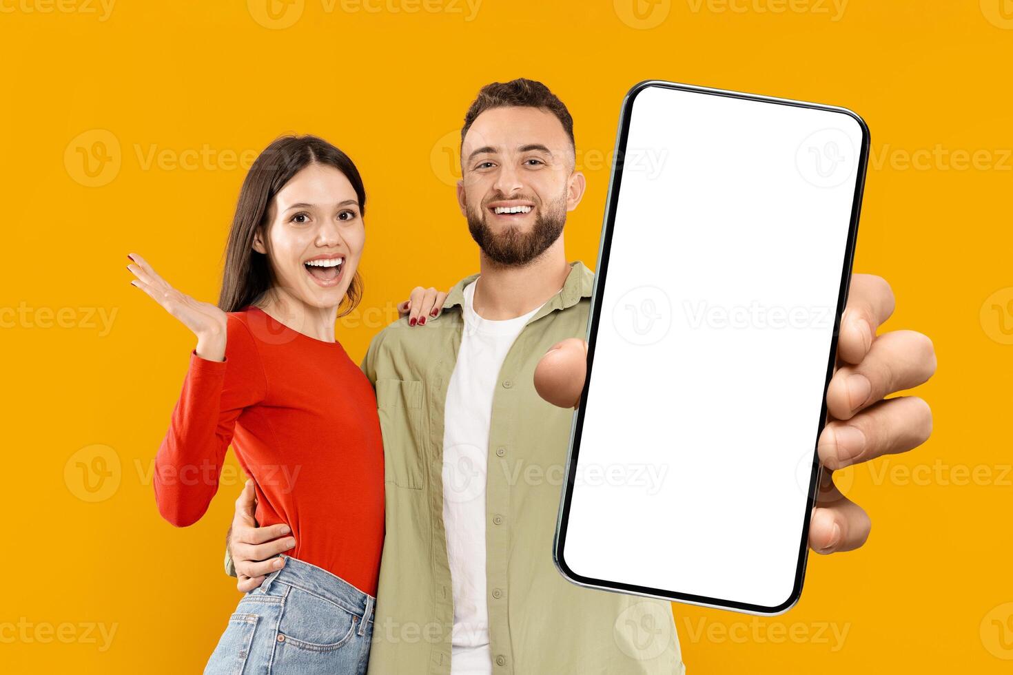 A young Caucasian couple enthusiastically poses with a smartphone in hand. The woman smiles widely, while the man grins happily, showcasing their love and affection. photo