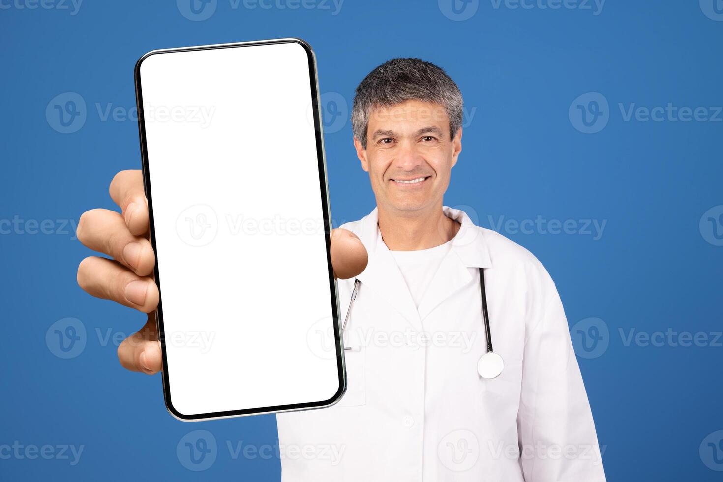 A mature Caucasian doctor in a white coat holds a blank smartphone in front of him. He is smiling, posing against a blue background, promoting digital health solutions. photo