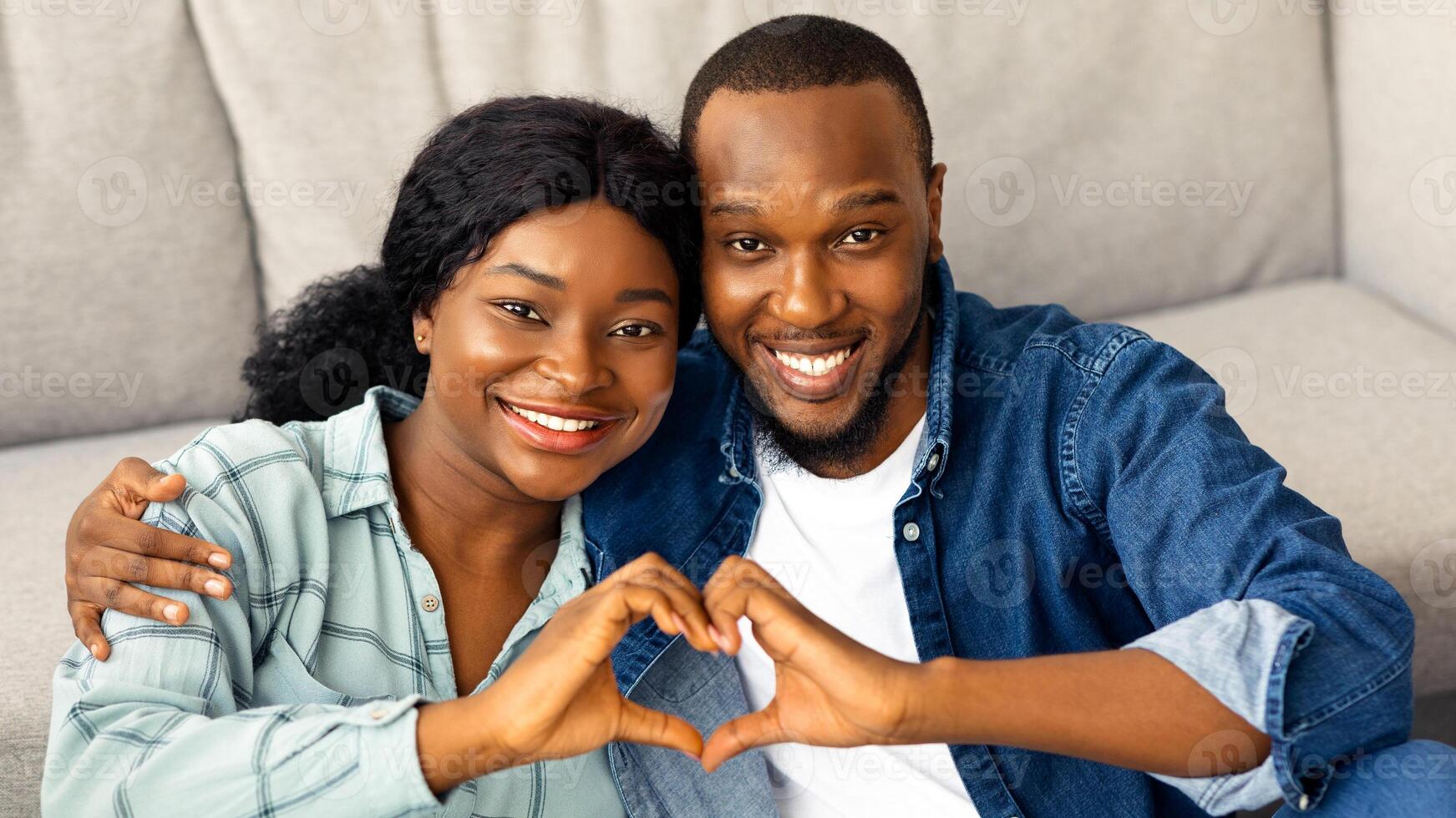 Portrait Of Joyful Romantic African American Couple Making Heart Gesture At Camera, Smiling Black Spouses Connecting Their Hands Together Into Love Sign While Relaxing At Home, Closeup Shot photo