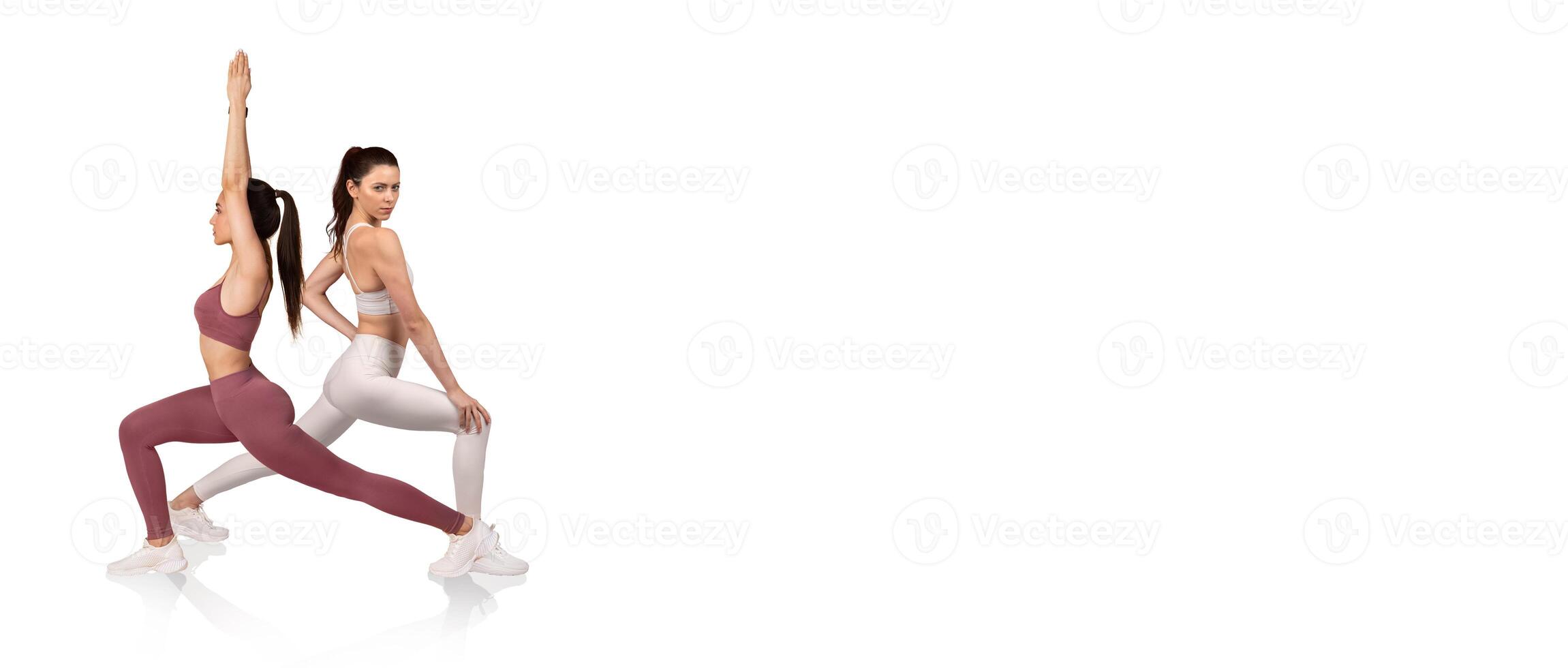 Two women perform yoga poses in a bright studio. One woman is in a warrior pose while the other is in a seated position, showcasing flexibility and strength in a peaceful setting. photo