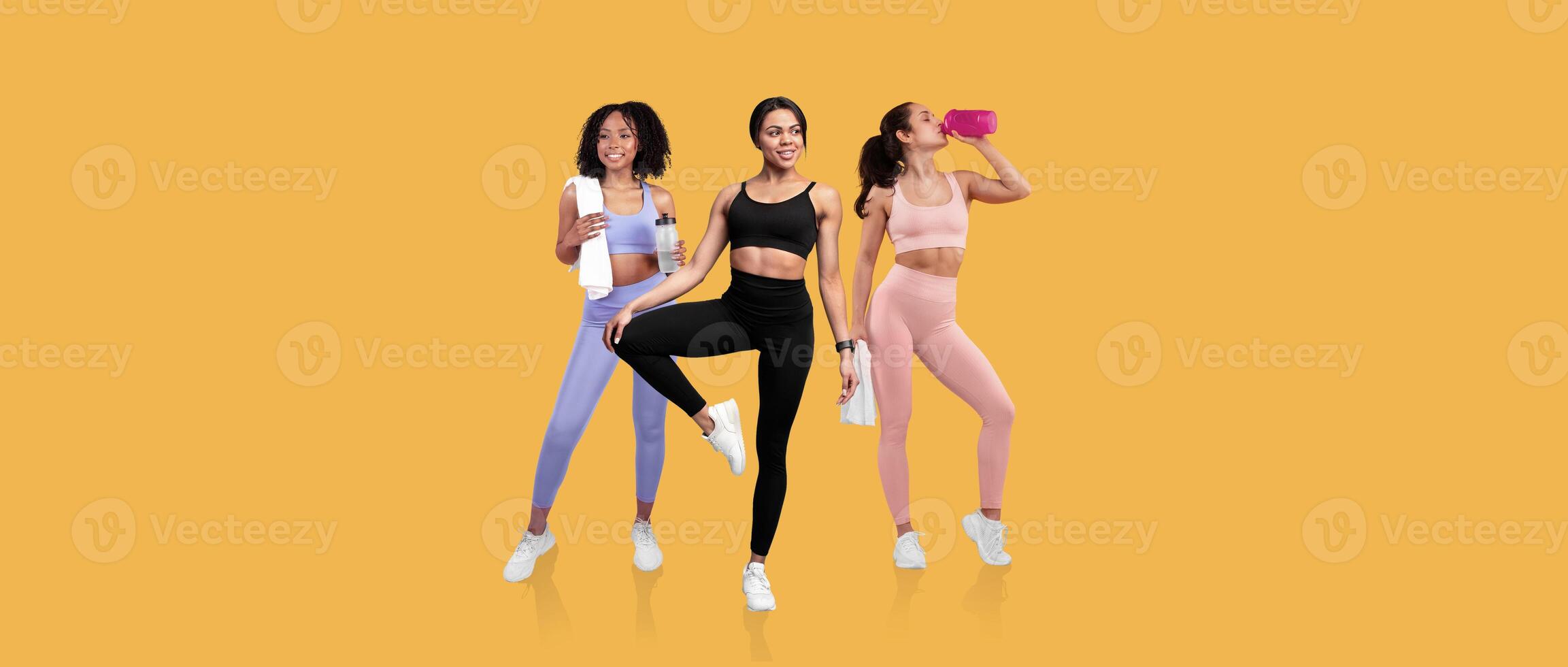 Three women of diverse backgrounds display fitness enthusiasm by stretching, posing, and hydrating in a vibrant setting. The bright yellow background adds energy and motivation to their workout. photo