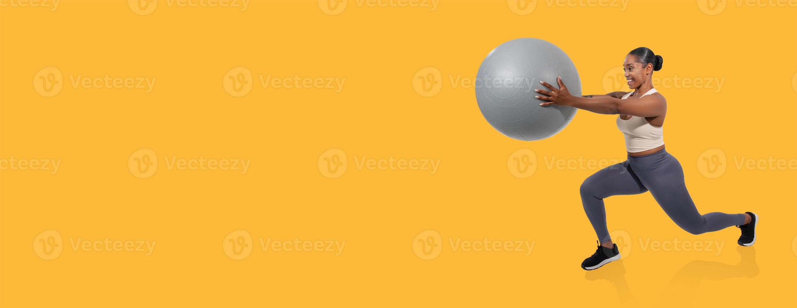A woman engages in a lunge exercise while holding a stability ball, demonstrating strength and balance. The bright yellow background adds energy to her workout routine. photo
