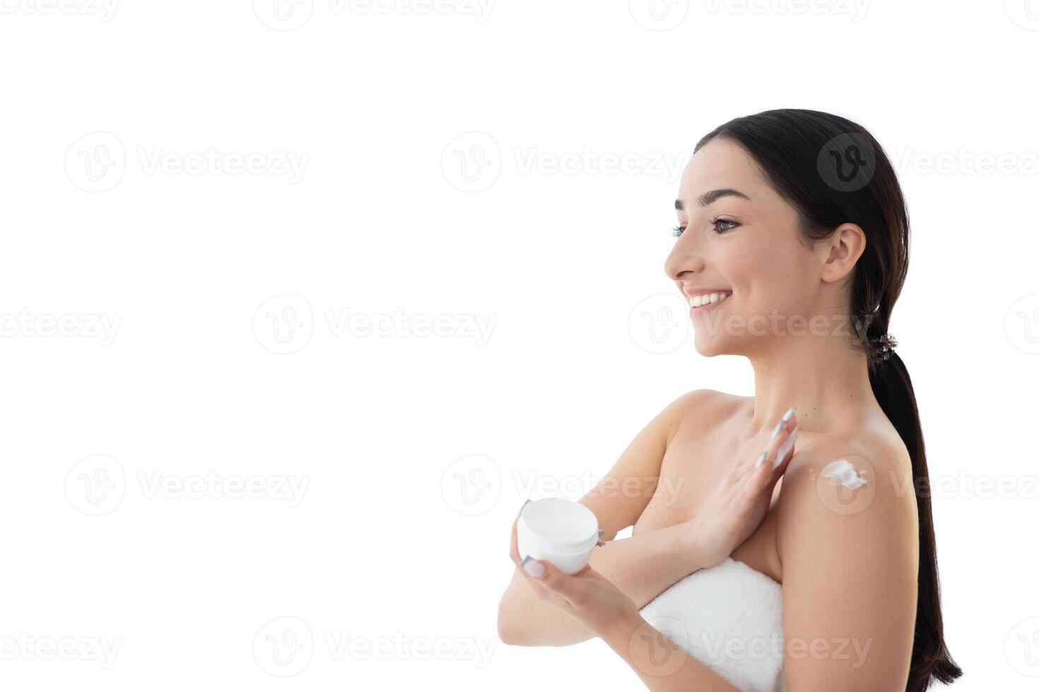 A young European woman enjoys some pampering, applying cream on her shoulder with a smile. She is in an isolated environment, focused on her beauty and skin care routine, copy space photo