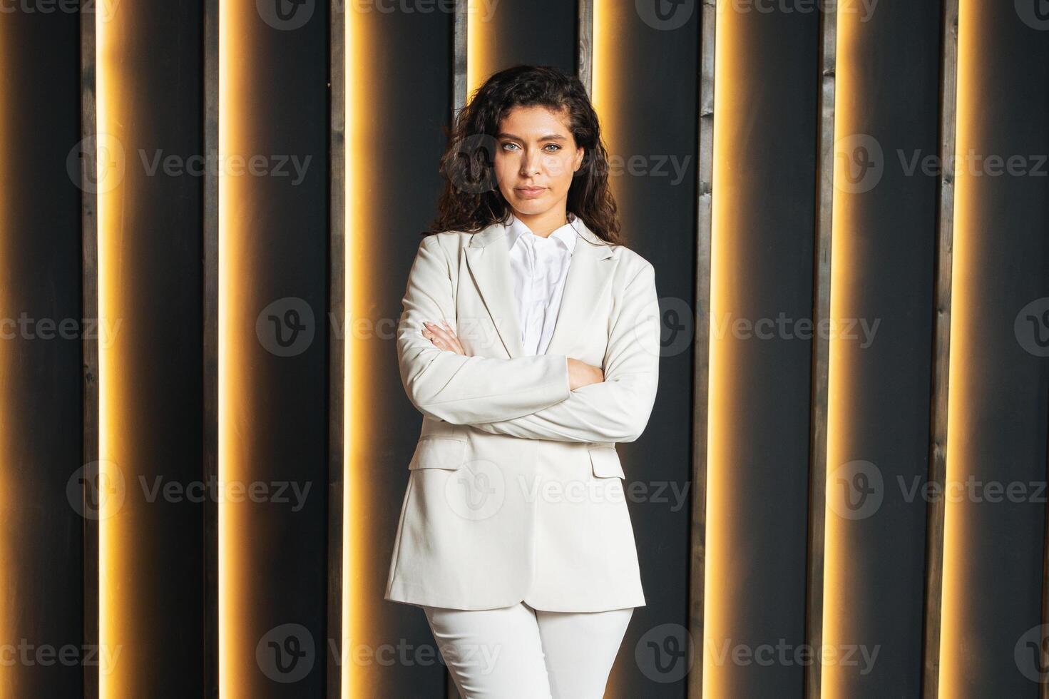 A European businesswoman stands confidently with crossed arms in a contemporary office setting. The backdrop features sleek lighting, emphasizing her professional demeanor and style. photo