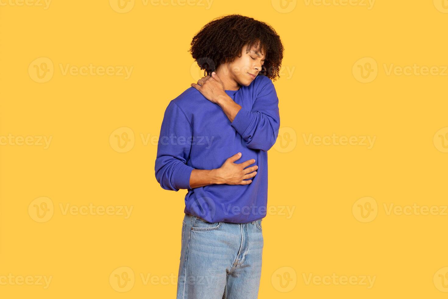 A man stands alone against a vibrant yellow background, holding his stomach and rubbing his shoulder with a pained expression. He showcases a relaxed yet hipster style in casual attire. photo