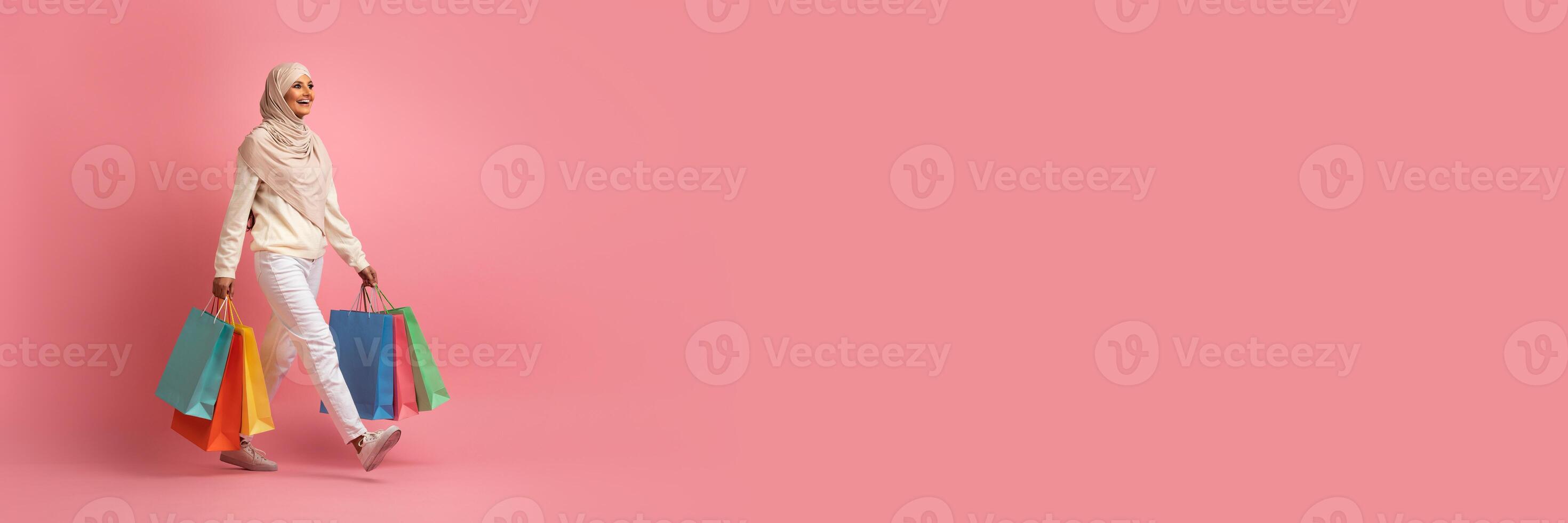 Muslim woman dressed in casual attire, carrying vibrant shopping bags, walks with a smile against a soft pink background. This moment captures the joy of shopping during seasonal sales, copy space photo