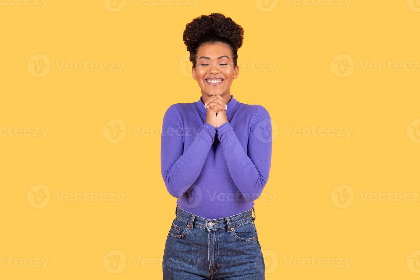 A young Hispanic woman with a joyful expression stands in front of a bright yellow background. She wears a purple top and denim jeans, embodying a cheerful lifestyle in an isolated setting. photo