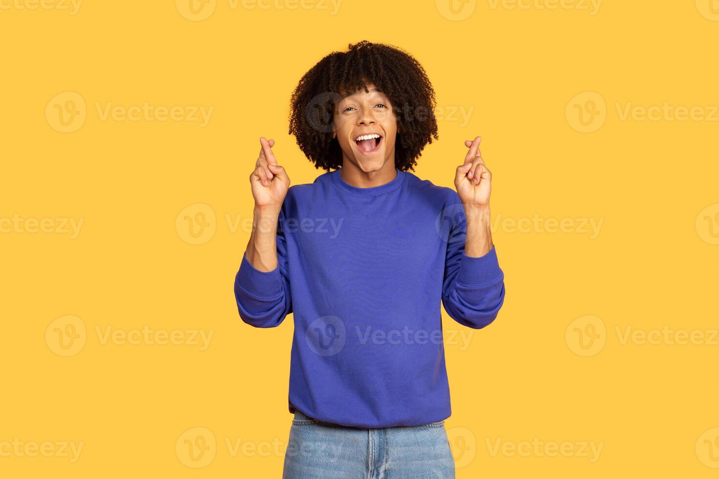 A young black man with curly hair stands against a vivid yellow background, smiling widely and playfully crossing his fingers, expressing hope and enthusiasm. It's a lively and cheerful moment. photo