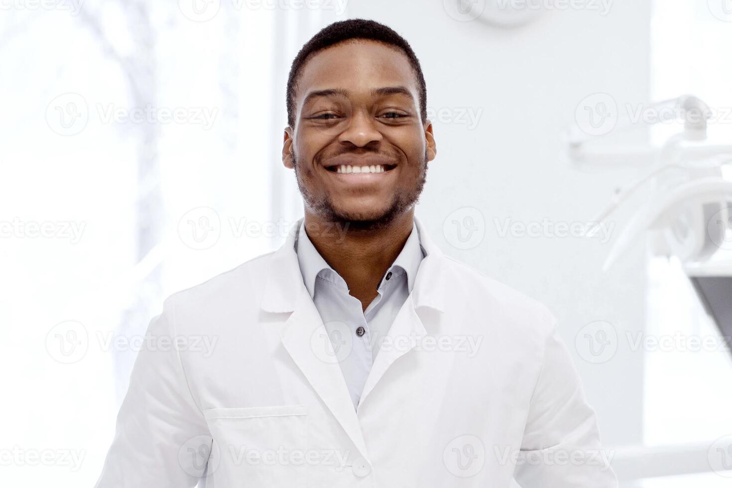 Portrait Of Handsome Young Black Dentist Doctor In Uniform Posing In Clinic. Professional Affican American Orthodont Standing In Modern Stomatological Cabinet And Smiling At Camera, Free Space photo