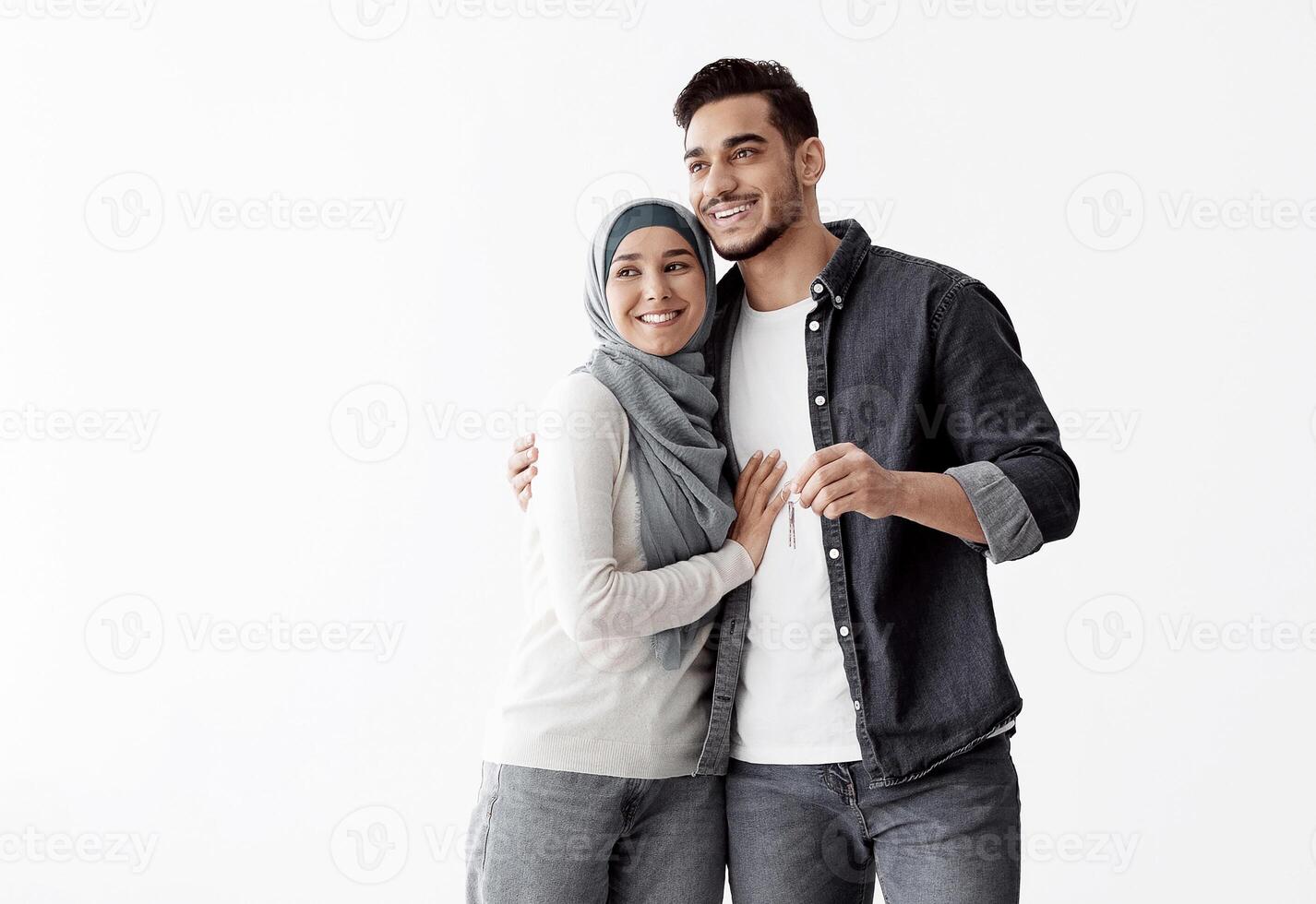 Happy young muslim couple with key standing in empty white room of their new apartment, hugging and looking at empty space for advertisement, panorama. Real estate, house buying cocnept photo