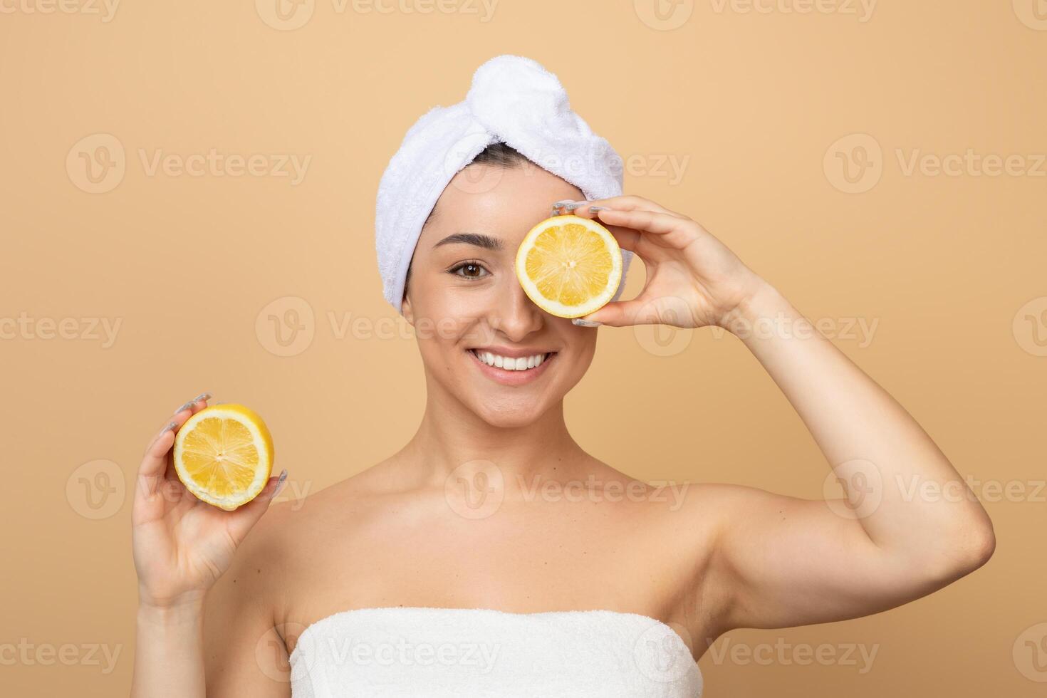 A young Caucasian woman is pampering herself with skincare, playfully holding lemon halves in front of her face. Her cheerful expression shines against a warm, neutral background. photo