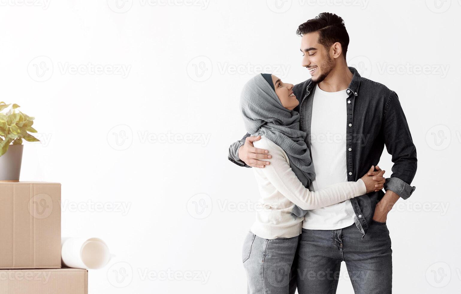 Loving muslim couple standing among moving supplies paper boxes with belongings in empty apartment, full length photo, copy space. Positive guy and young lady in hijab relocating photo