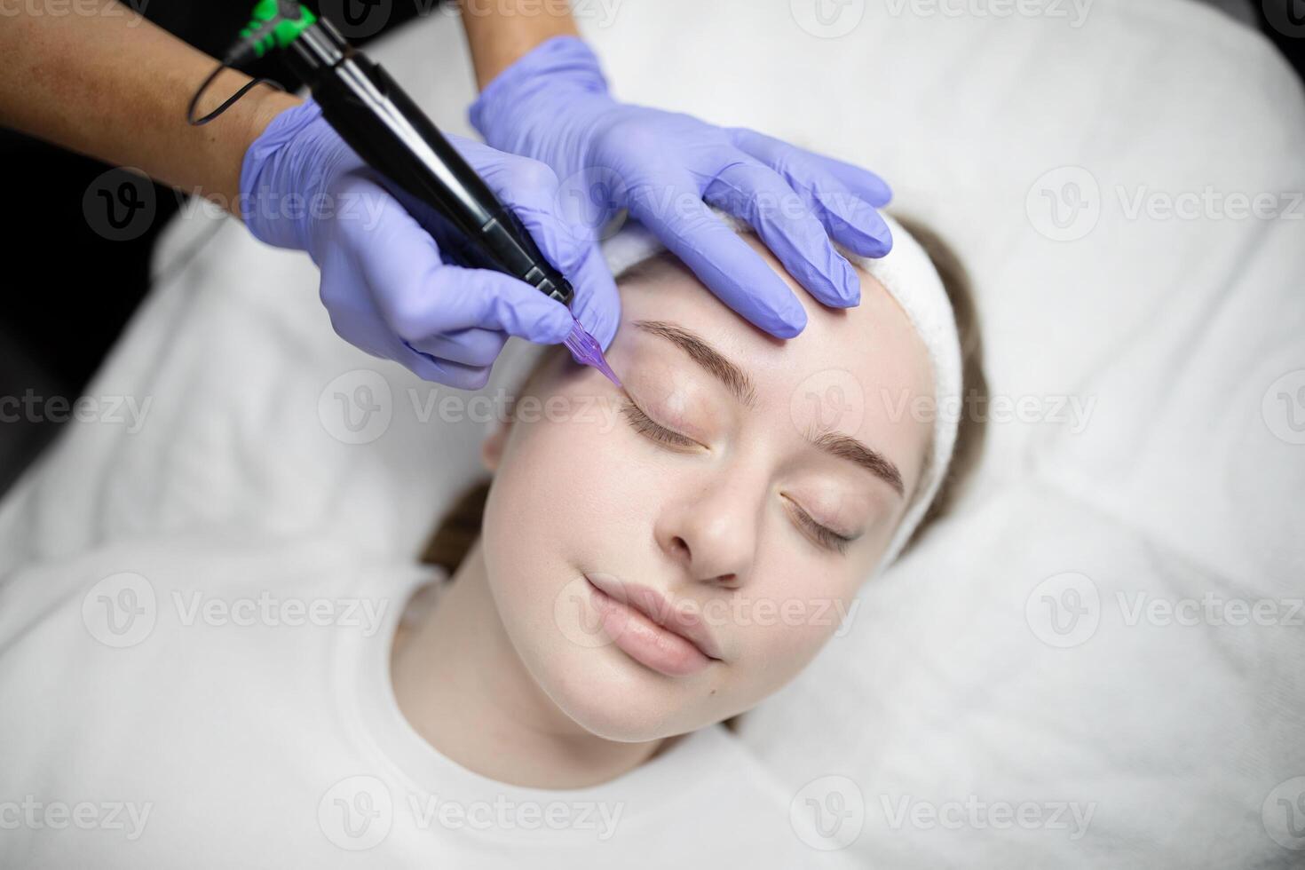 A woman relaxes while receiving a long-lasting makeup treatment at a beauty salon. The skilled technician uses a device for precise application, enhancing her features for a polished look. photo
