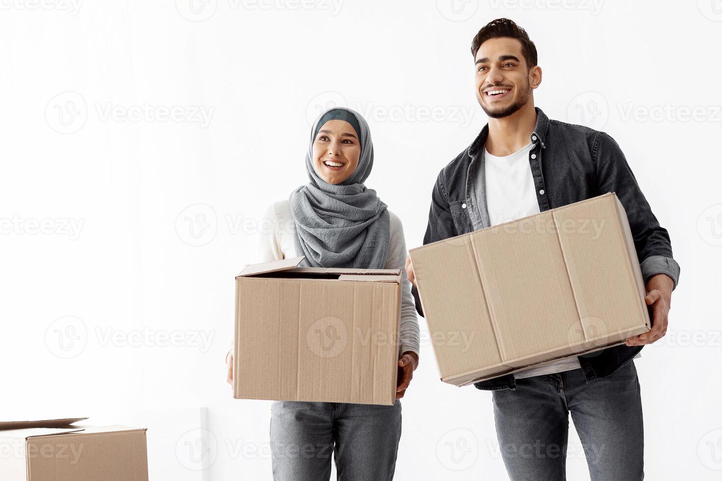 Happy young muslim couple moving to new apartment, standing together over white wall, holding big paper boxes full of their belongings, looking aroung and smiling, copy space, full length shot photo