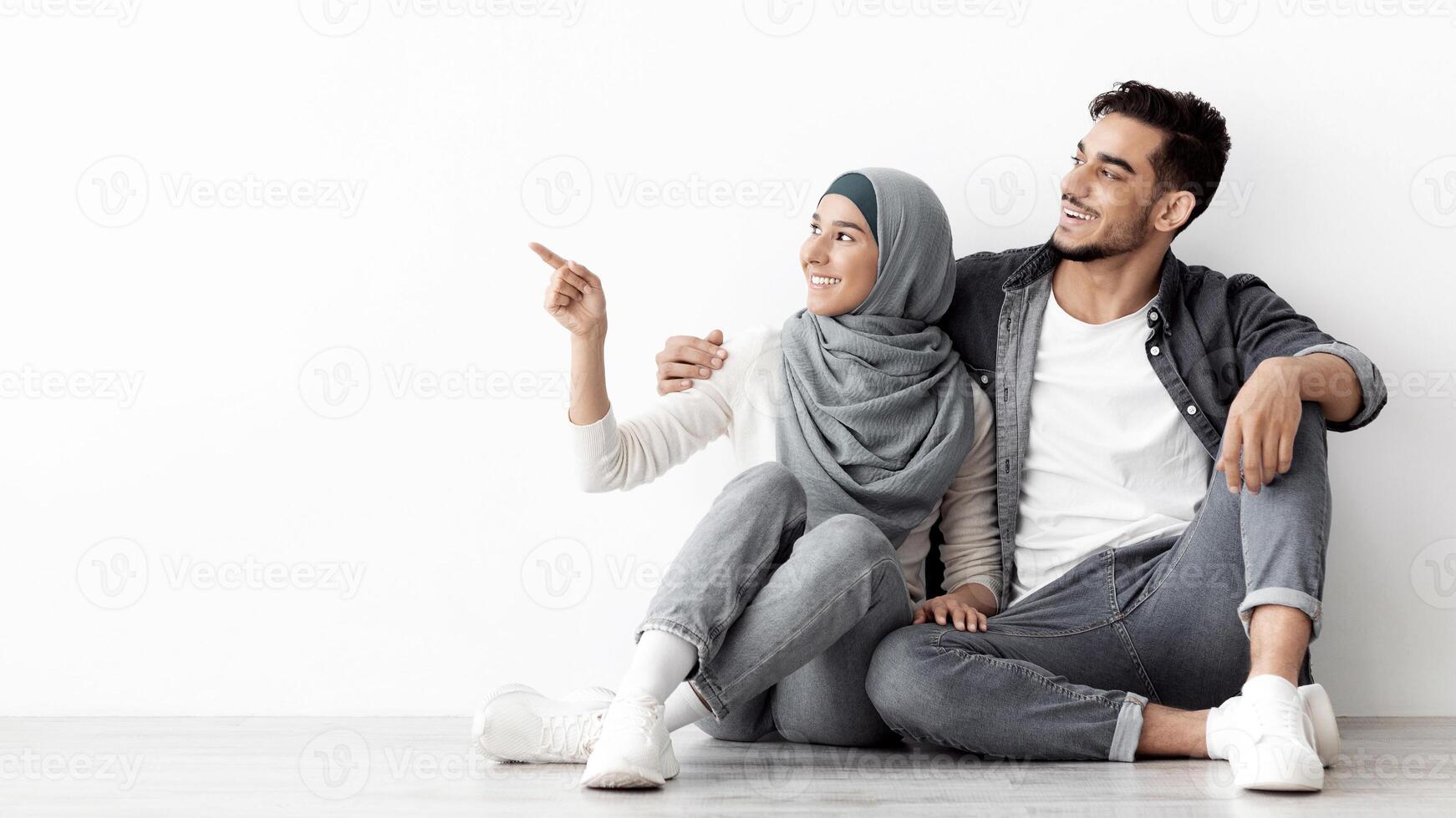 Dreamy young woman in hijab sitting with her loving husband on floor in new empty apartment, pointing at copy space for advertisement over white wall, embracing and smiling, panorama photo