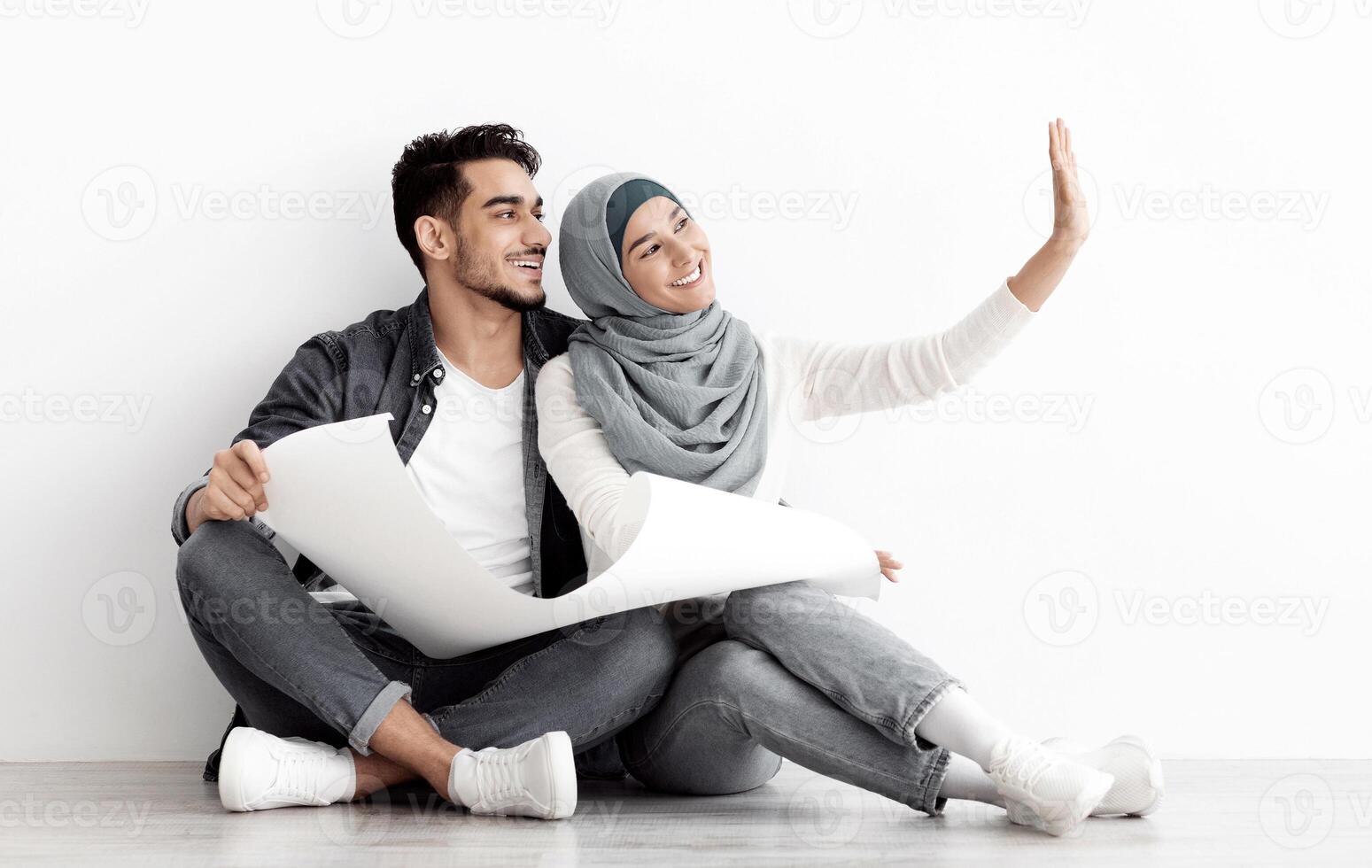 Emotional muslim couple thinking about design in their new house, sitting on floor over white empty wall, holding apartment plan and imaging future interior, panorama with copy space photo