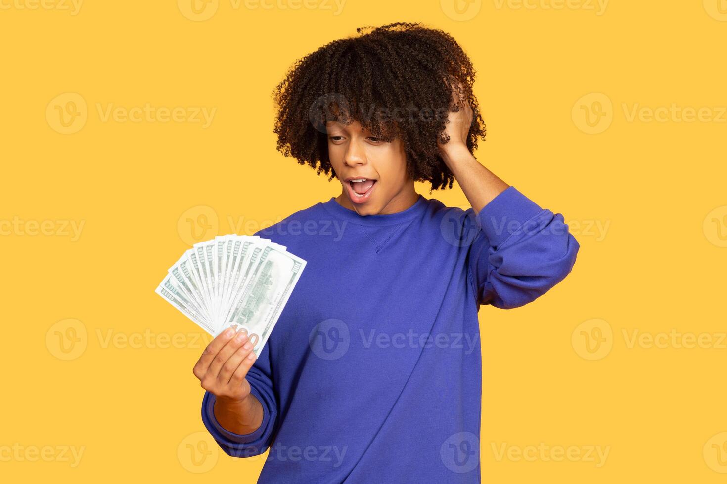 A young Brazilian man with curly hair looks surprised while holding a fan of bills in his hands. The vibrant yellow background enhances his joyful expression and playful mood. photo