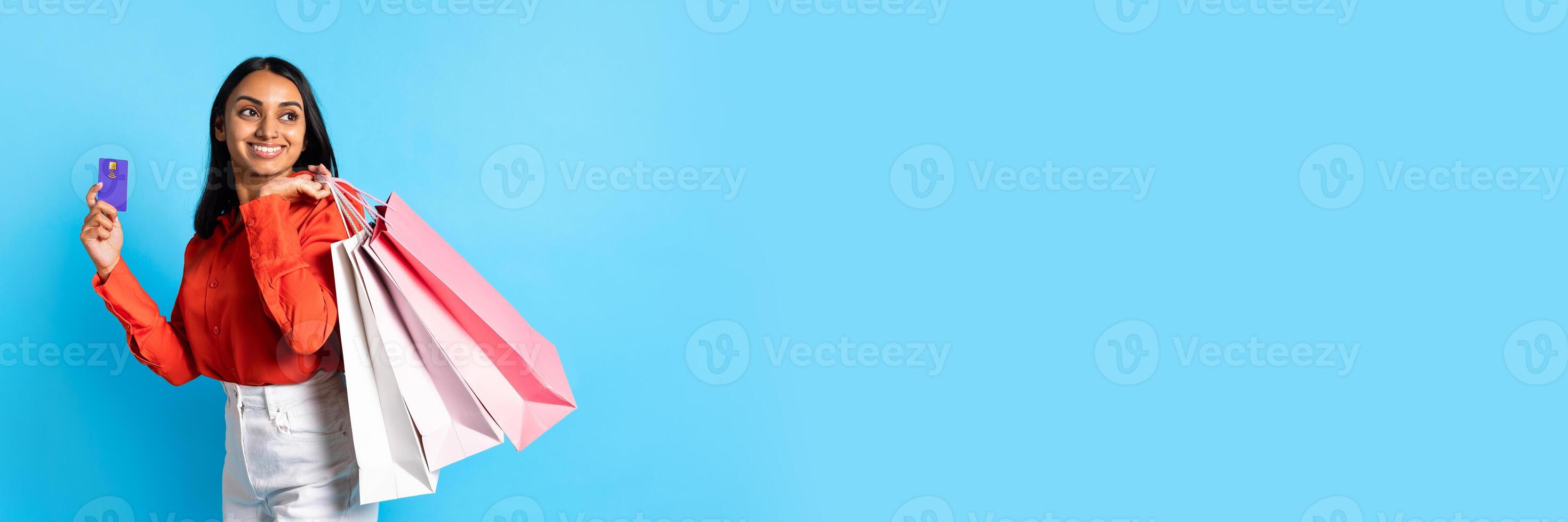 Shopping Offer. Happy Young Indian Woman In Casual Holding Credit Card And Shopper Bags, Standing On Blue Studio Background. Banner For Advertising Sale Season At Mall. Panorama With Copy Space photo