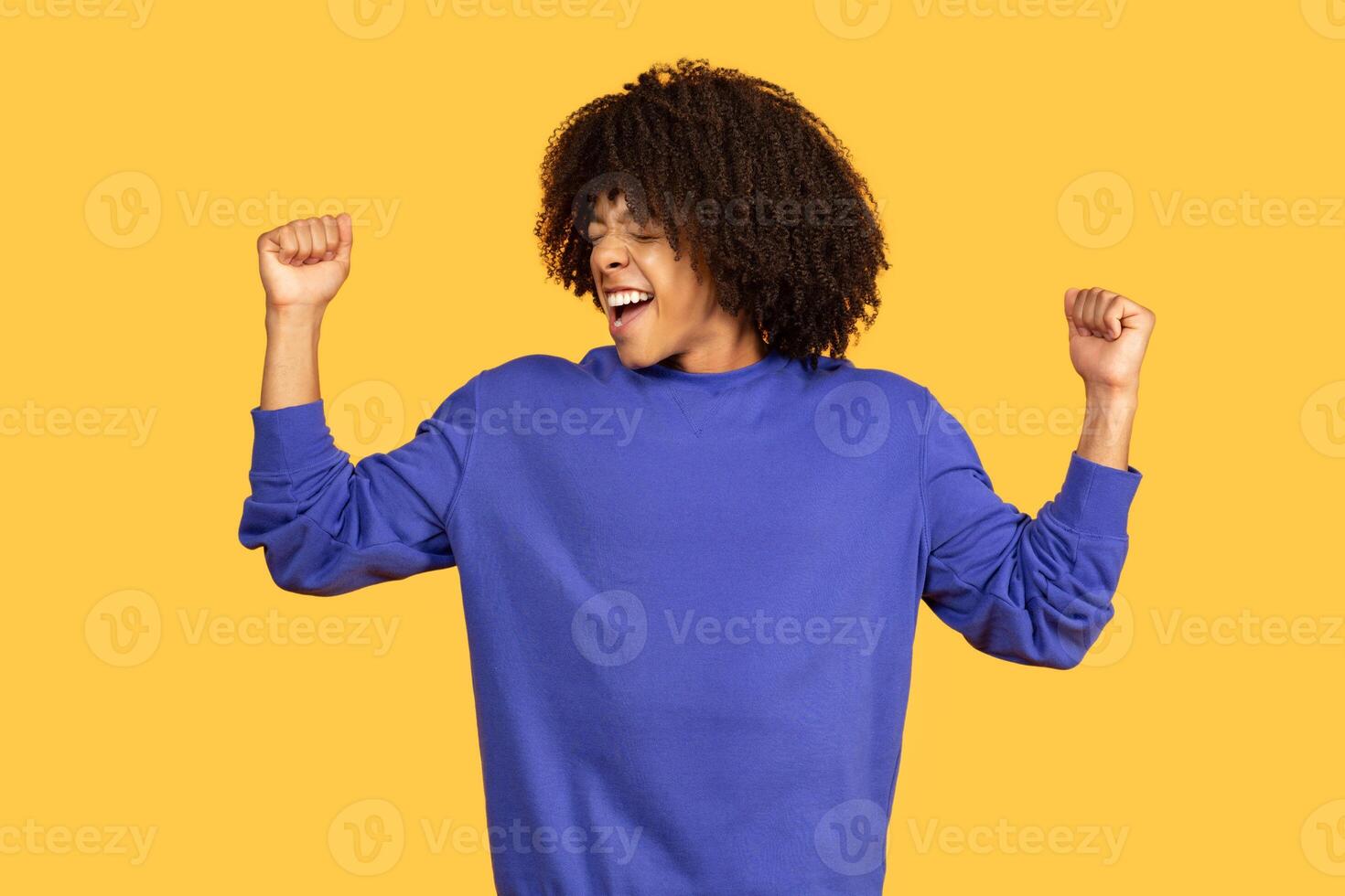 A young black man with curly hair is joyfully celebrating with raised arms in front of a bright yellow backdrop. His expression conveys excitement and energy, representing a modern lifestyle. photo