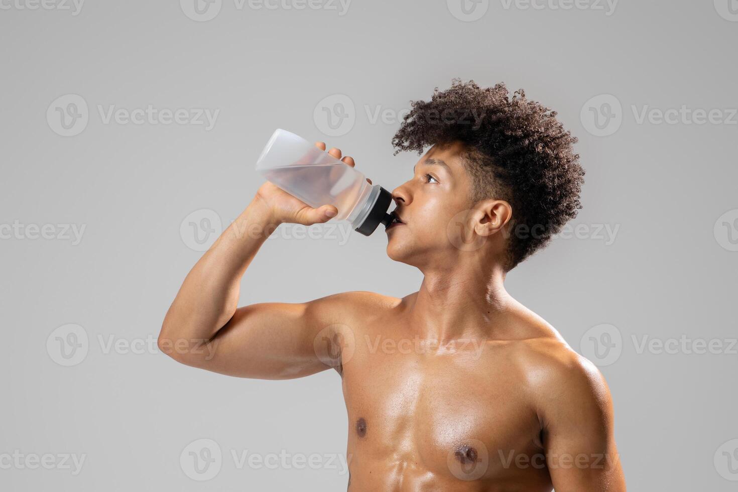 A young Brazilian man with an athletic build is quenching his thirst with a water bottle. He is in a gym setting, highlighting a commitment to a healthy lifestyle and fitness. photo