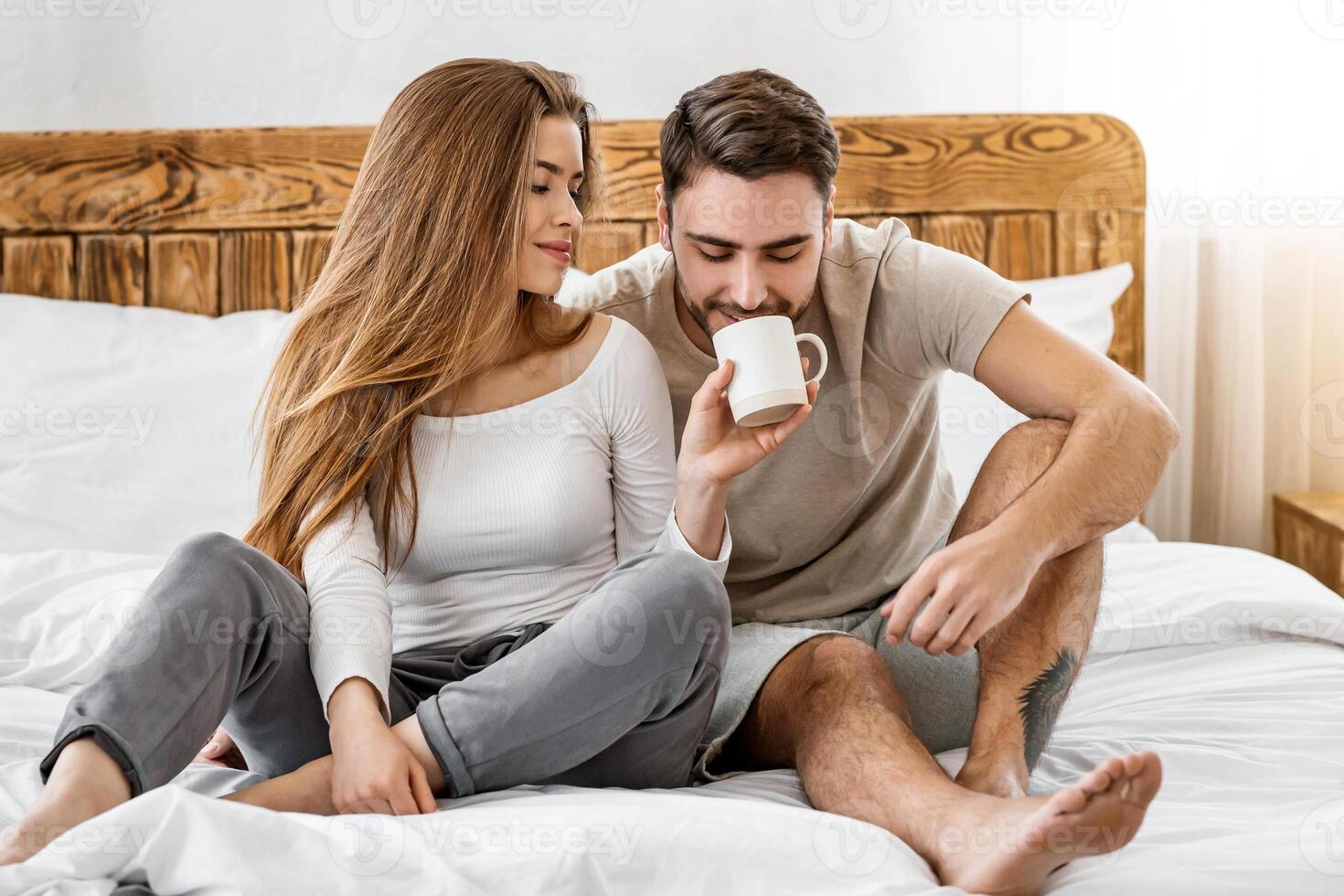 Romantic coffee in bed for two. Smiling young woman gives her husband drink from cup in interior of bedroom, panorama, free space photo