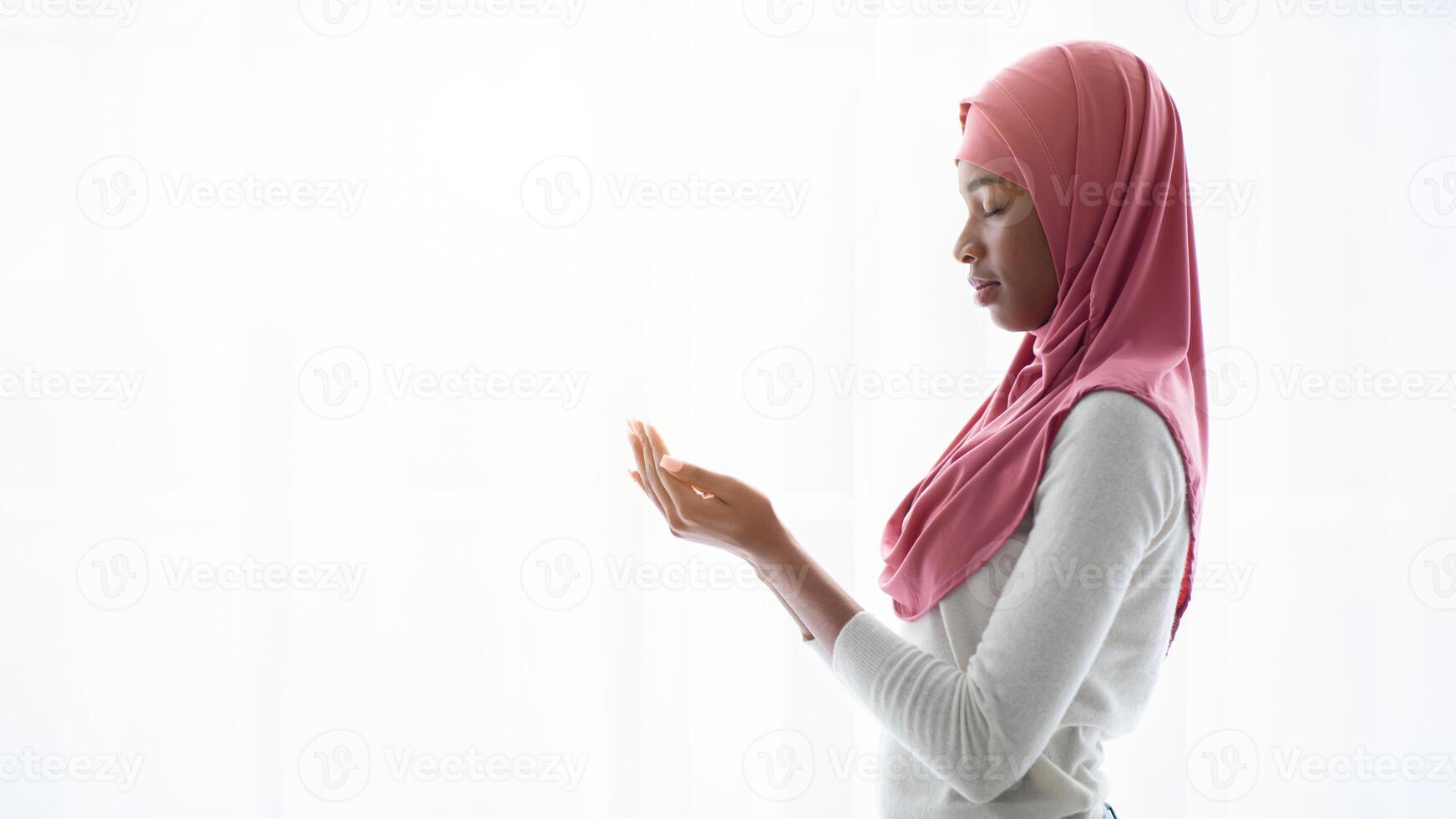 Portrait of young black muslim woman praying near window at home, religious lady in headscarf standing with closed eyes, making islamic pray dua gesture with cupped hands, side view, copy space photo