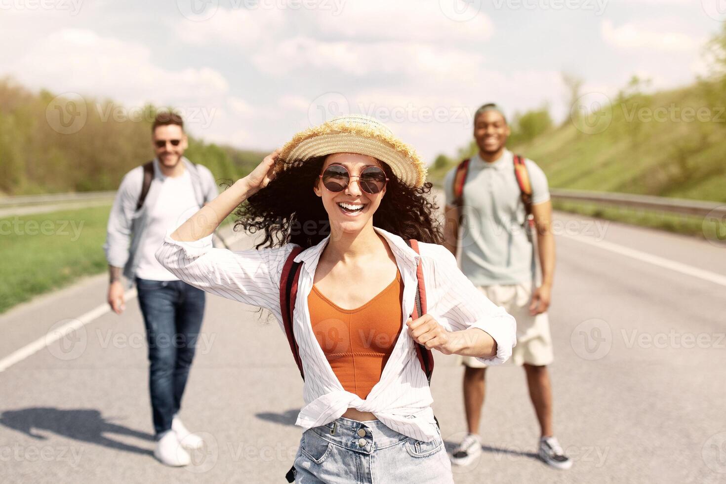 Beautiful young woman walking along highway with her male friends, having summertime journey, backpacking outdoors. Hitchhiking, autostop travel, summer adventure and tourism concept photo