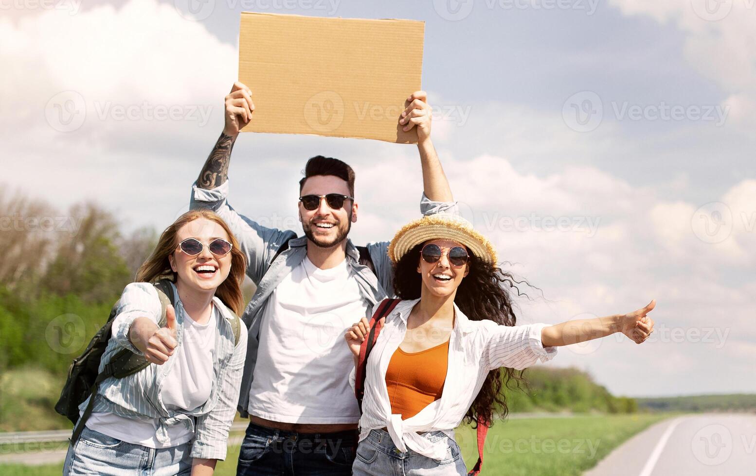 Cheerful diverse friends with backpacks and guitar hitchhiking on highway, catching free ride, having summer adventure, copy space. Autostop journey, summertime freedom, travel and friendship concept photo