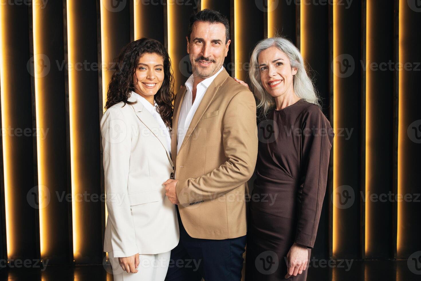 A diverse group of executives, including a man and two women, stands together in a modern office. They engage in a collaborative discussion, showcasing a professional atmosphere and teamwork. photo