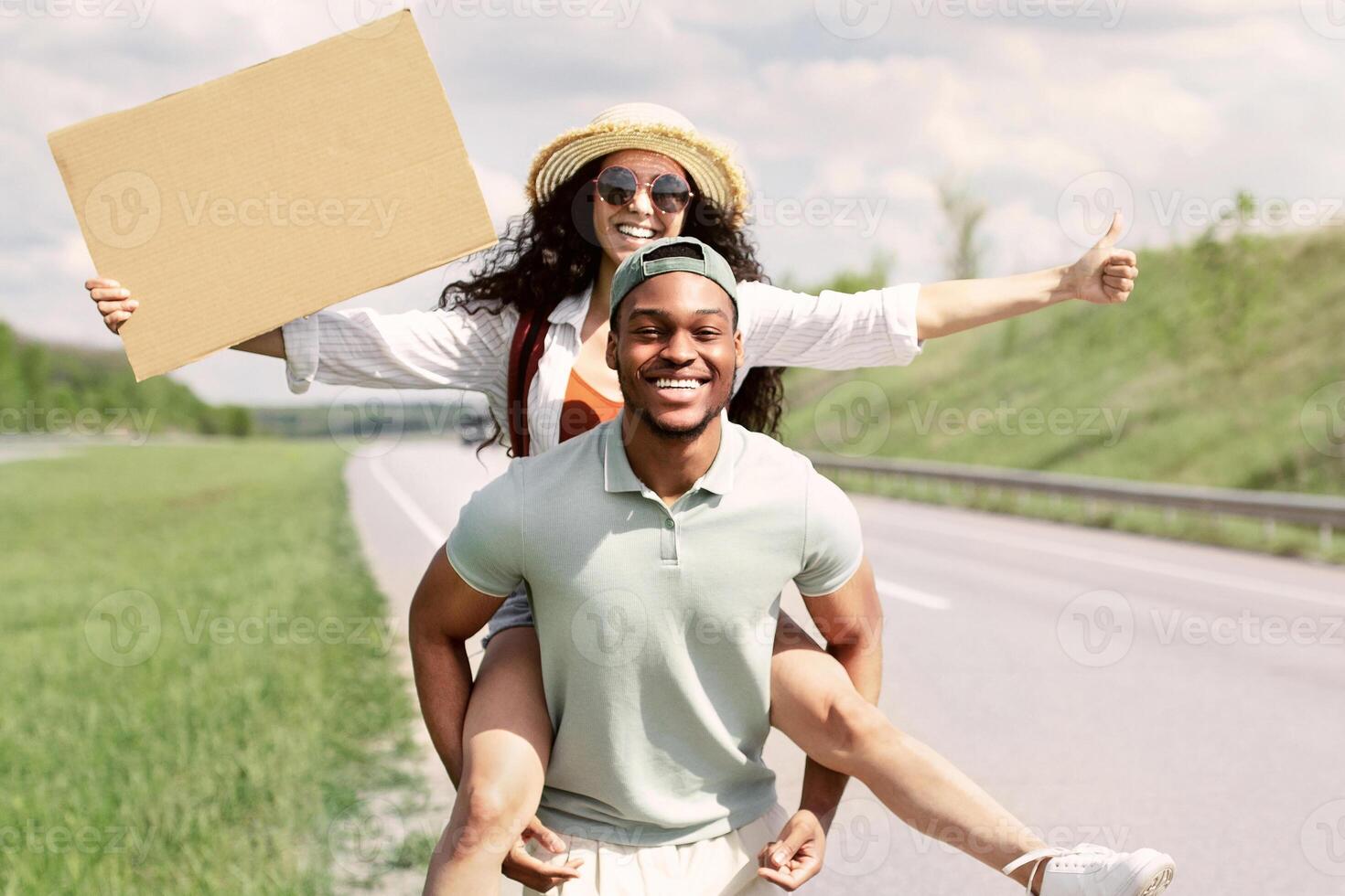 Full length of happy multiracial couple hitchhiking, trying to stop car with empty cardboard sign, mockup. Black guy giving his girlfriend piggyback ride on highway. Autostop travel, summer vacation photo