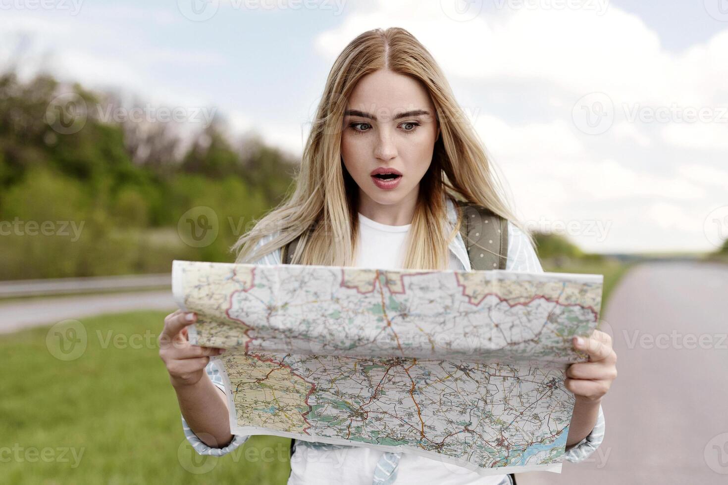 Shocked young woman hitchhiking on road, looking at map in terror, feeling lost while traveling alone by autostop. Millennial blonde lady standing on highway, cannot find her destination photo