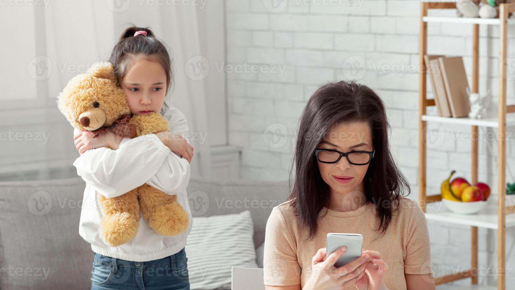 Lonely teen girl hugging teddy bear while her busy mom working online from home, not paying attention to child. Depressed kid feeling neglected, missing her parent. Covid-19 quarantine family problems photo