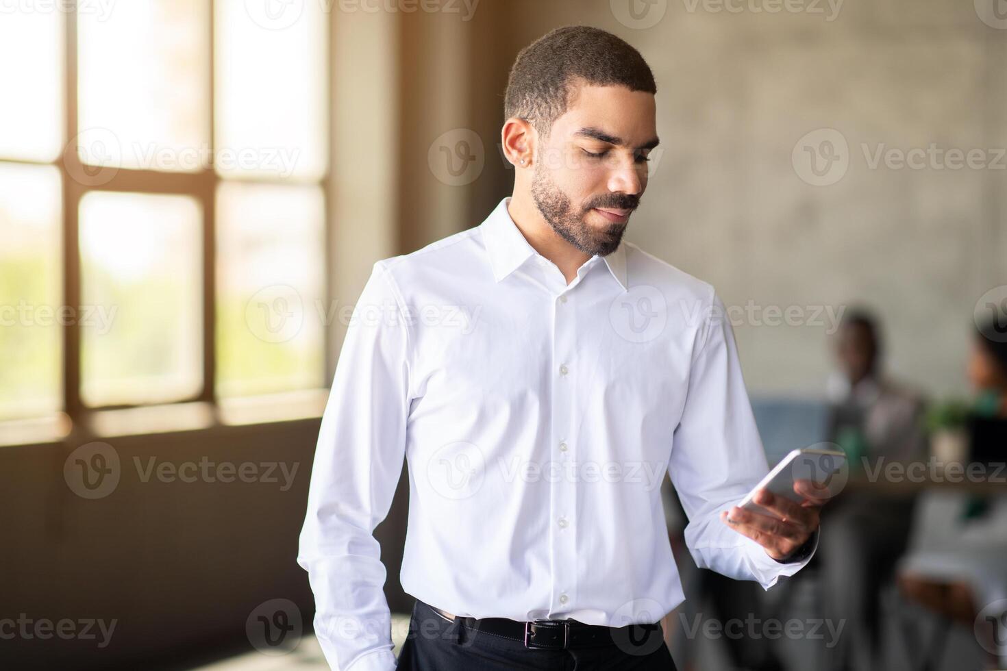 A business professional checks his smartphone in a contemporary office environment. The warm lighting and diverse team in the background enhance the atmosphere of collaboration and productivity. photo