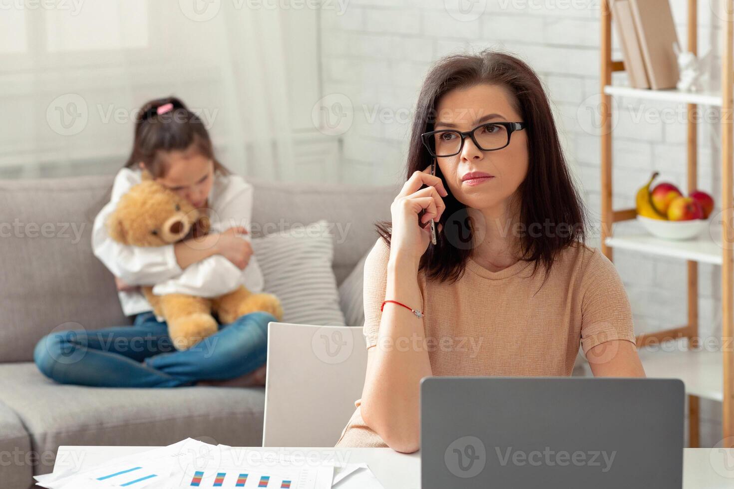 Modern working mom doing online job, her neglected daughter hugging teddy bear, feeling lonely and sad at home. Young mother working remotely during covid-19 pandemic, ignoring her child photo
