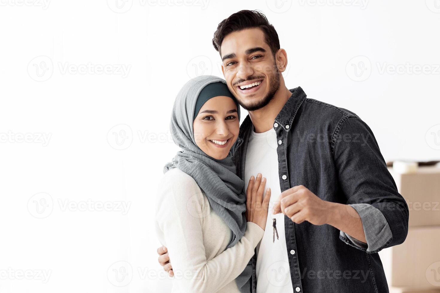 Happy muslim family standing over paper boxes and showing keys from their new apartment. Handsome arab man and young woman in hijab moving to their house, unpacking belongings, copy space photo