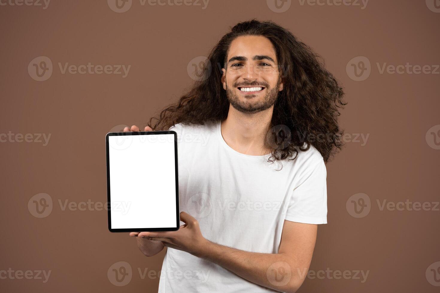 A young Caucasian man with long curly hair smiles brightly while holding a blank tablet. He stands against a simple brown background, showcasing a relaxed and positive lifestyle. photo