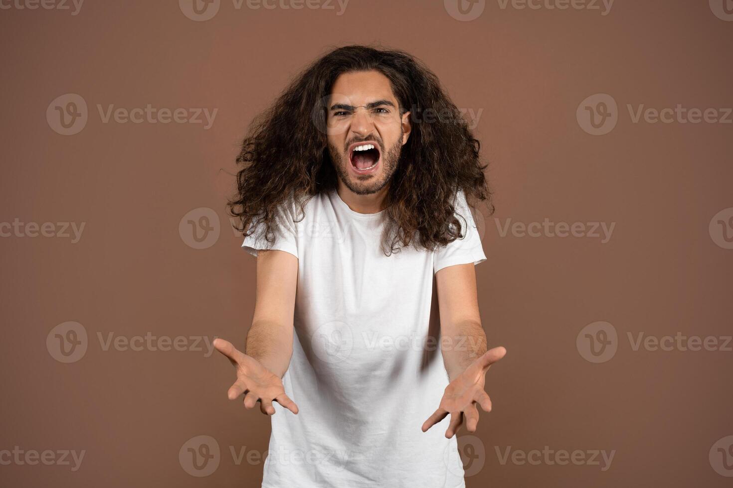 A young European man with curly, long hair appears passionately expressive while standing in a studio. The neutral background contrasts with his vibrant energy and animated gestures. photo