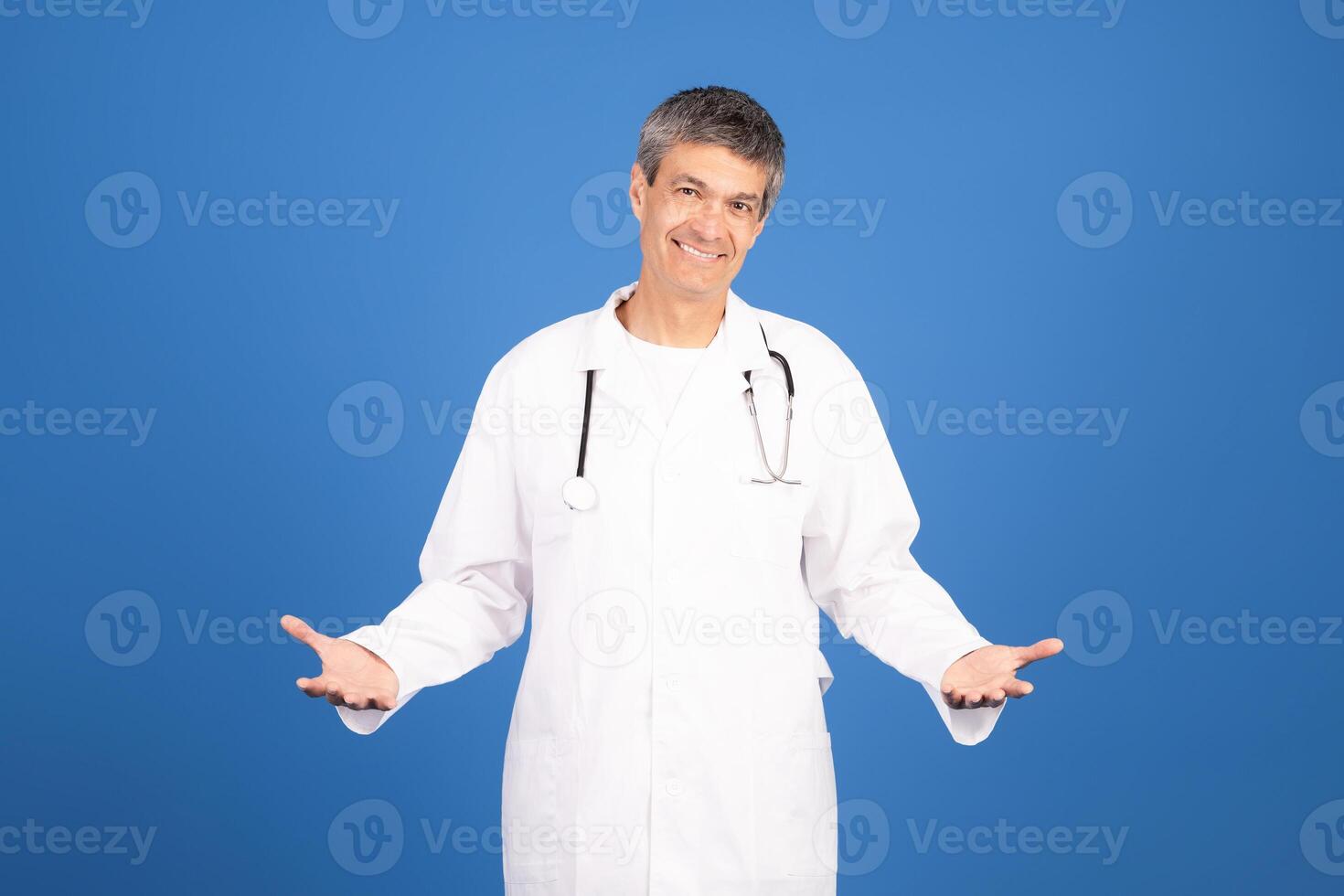 A mature Caucasian man in a white doctor coat stands confidently with a welcoming smile. He gestures with open arms, promoting a friendly atmosphere in a healthcare setting. photo