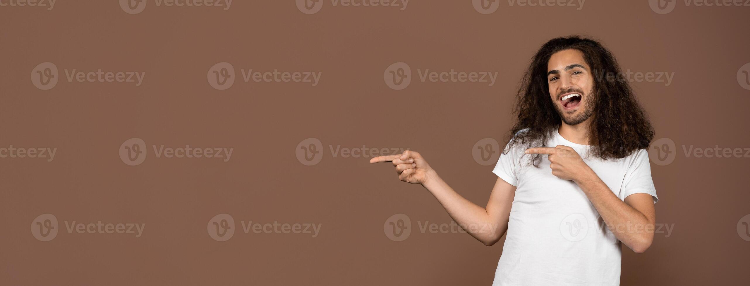 A young European man with curly hair joyfully points to copy space in a minimalist environment. His casual t-shirt and spirited demeanor convey a relaxed, trendy lifestyle. photo