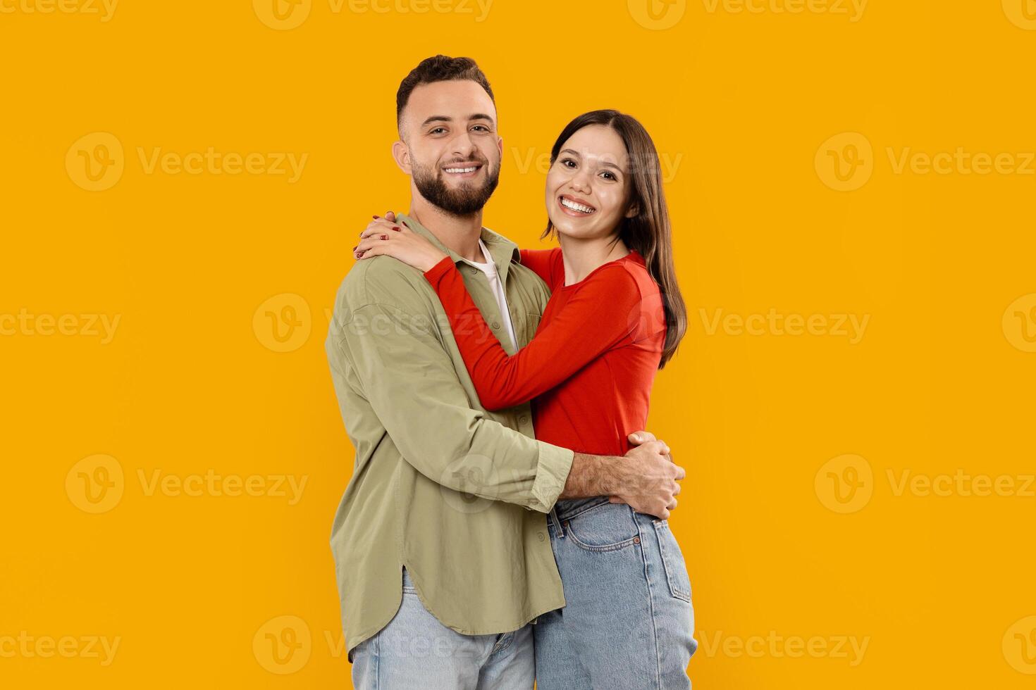 A married couple stands closely together, smiling joyfully. The man wears a light green shirt, while the woman is dressed in a vibrant red top. Their warm embrace showcases their affection. photo