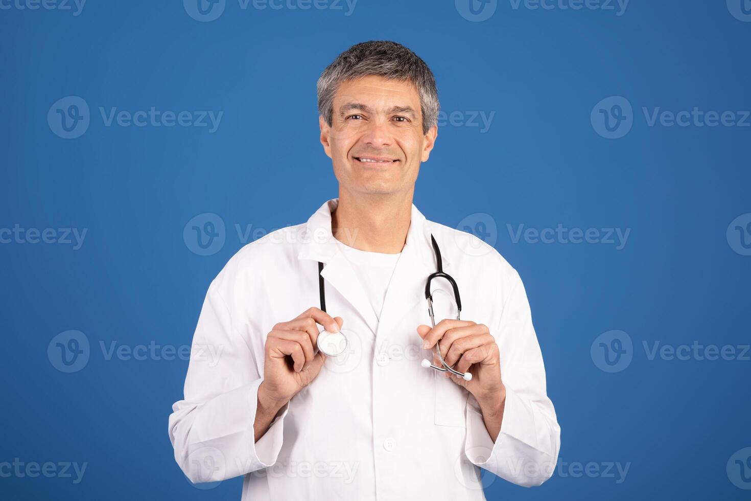 A mature European man in his 50s, dressed in a white coat, smiles confidently while holding a stethoscope. The background is a plain blue, emphasizing his role in healthcare and general medicine. photo