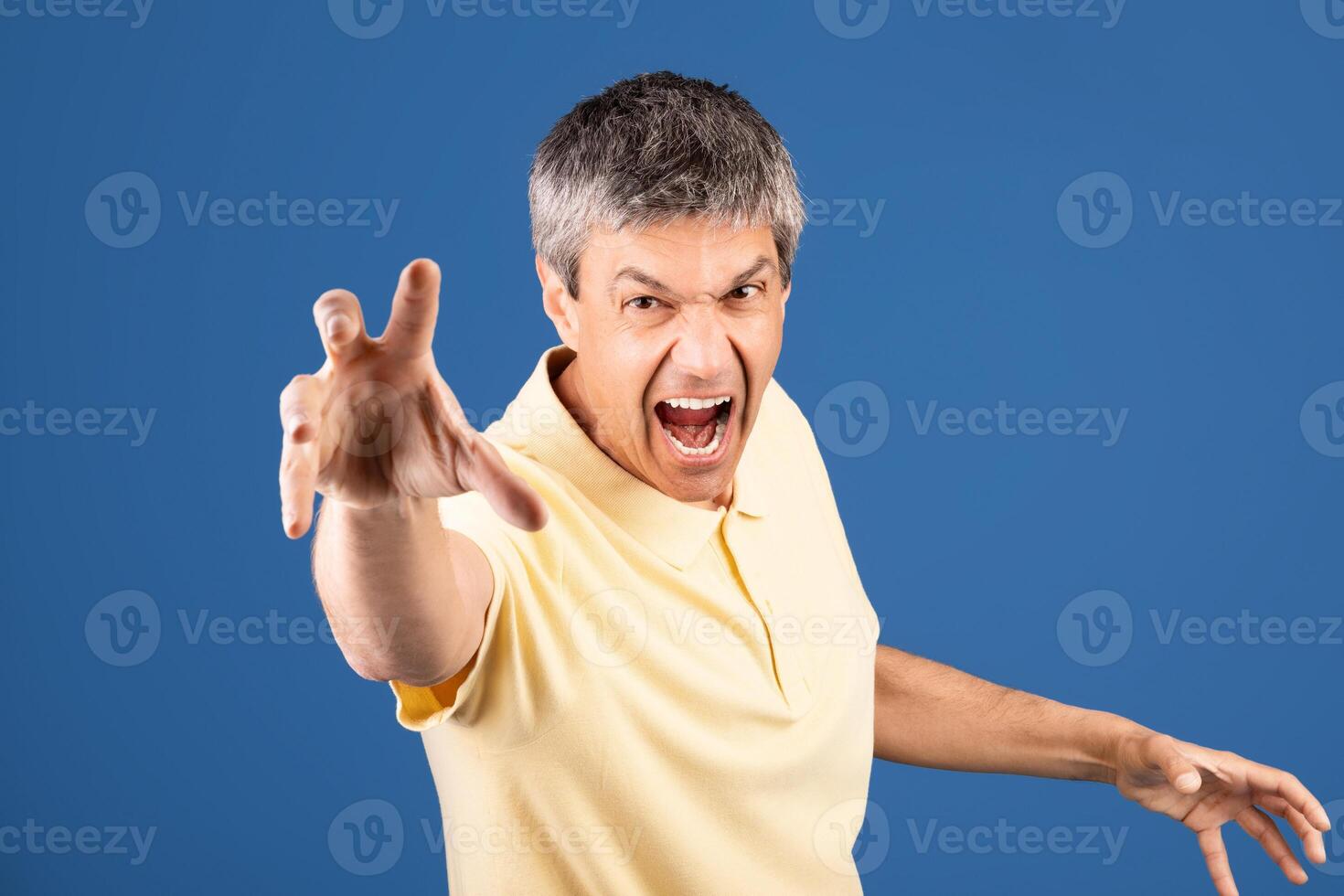 A middle aged man in his 50s is passionately expressing intense emotion. He appears to be in a studio with a blue background, wearing a yellow shirt and with his hand extended forward. photo