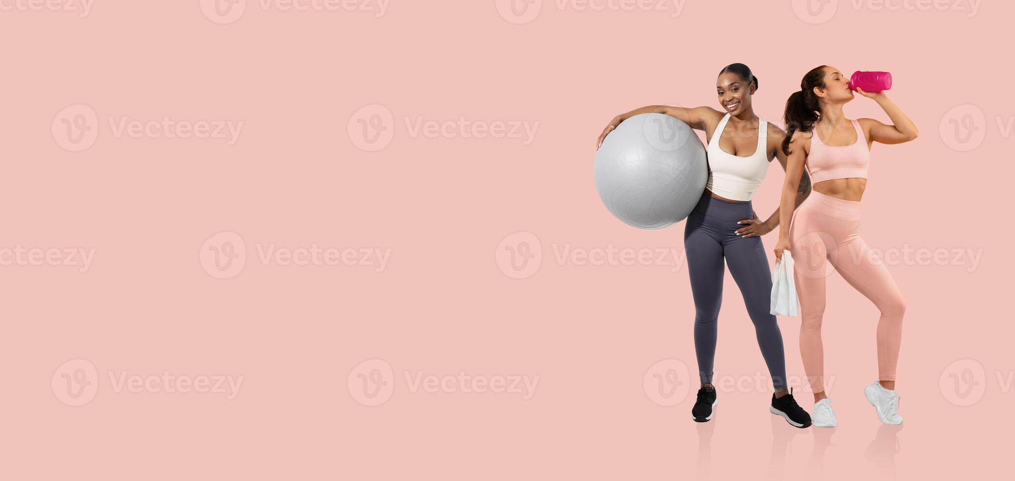 Two women are engaged in an active workout session in a fitness studio. One holds a large exercise ball, while the other drinks from a sports bottle, showcasing a healthy lifestyle. photo