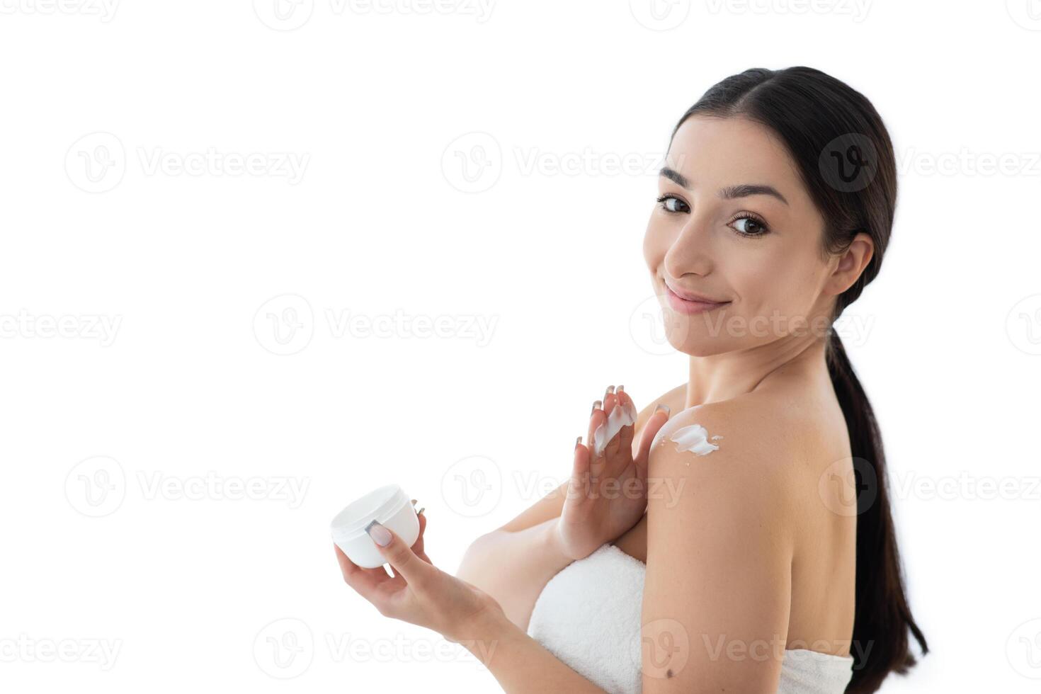 A young Caucasian woman applies cream to her shoulder, smiling as she indulges in her skincare routine. The minimalist setting highlights her focus on self-care and beauty, copy space photo