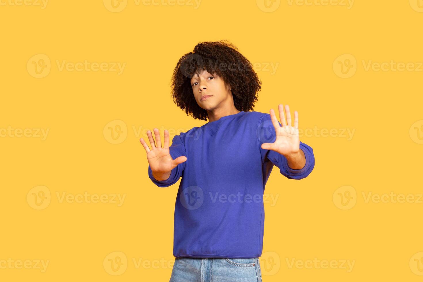 A Brazilian guy stands against a bright yellow backdrop, wearing a blue sweater and jeans. He strikes a playful pose with arms raised, showcasing a confident and relaxed vibe. photo