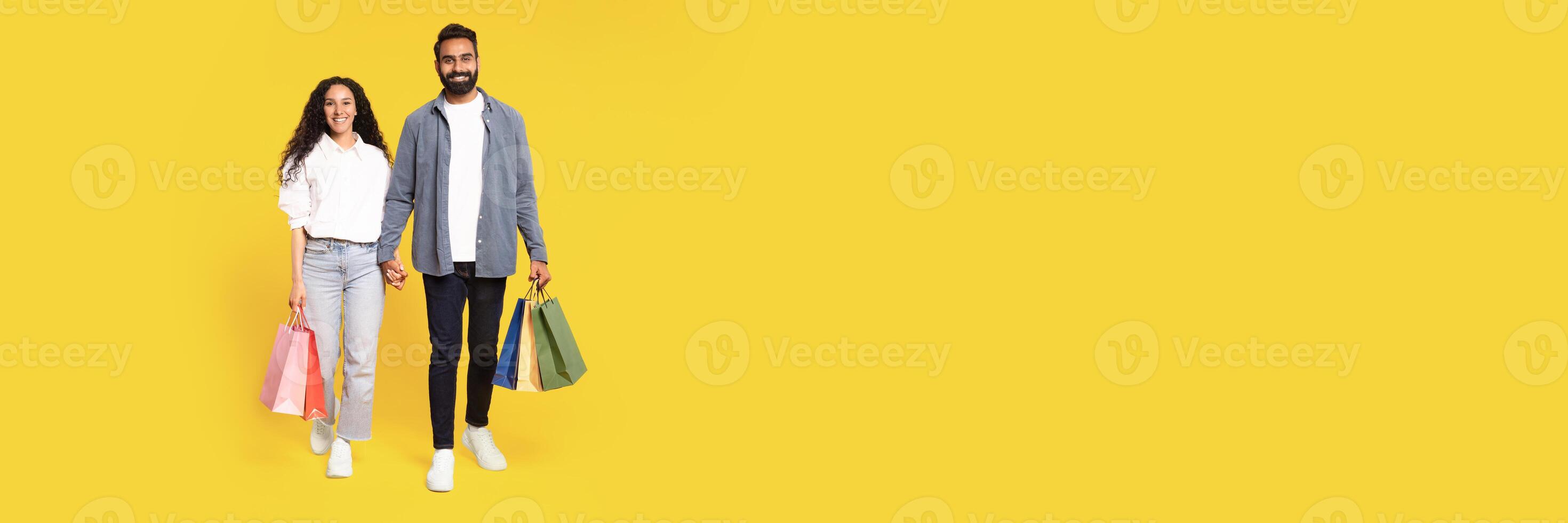 Big Sales. Full Length Of Happy Arab Couple Walking With Shopping Bags In Studio On Yellow Background. Smiling Young Middle Eastern Man And Woman Holding Hands Carrying Purchases, Copy Space photo
